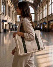 Load image into Gallery viewer, A woman walking through a large, sunlit train station hall carrying a cream canvas tote bag with dark green straps and a small white logo print.
