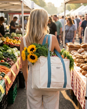 Load image into Gallery viewer, A woman at a farmers market carries a large off-white canvas tote bag with blue straps, filled with sunflowers and vegetables, walking between market stalls.
