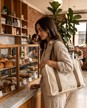 Load image into Gallery viewer, A woman in a beige linen outfit stands at a bakery counter in a cafe, carrying a large beige canvas tote bag over her shoulder.
